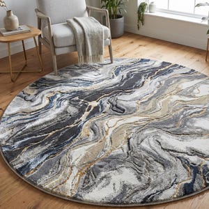 May include: A round area rug with a marbled design in shades of gray, white, and blue, accented with gold lines. The rug is placed on a wooden floor, with a chair and side table in the background. The rug's diameter is approximately 7 feet.