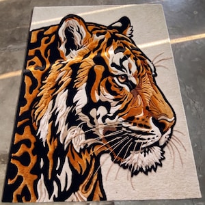 K&ouml;nnte beinhalten: Ein rechteckiger Teppich mit einem detaillierten Tigerkopf-Design. Der Teppich zeigt eine Profilansicht des Gesichts des Tigers mit orangefarbenem, schwarzem und wei&szlig;em Fell. Der Hintergrund des Teppichs ist eine neutrale beige Farbe, und die Schnurrhaare des Tigers sind sichtbar.