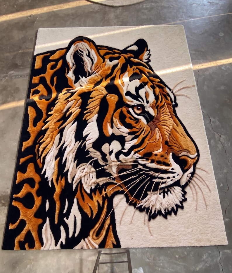 K&ouml;nnte beinhalten: Ein rechteckiger Teppich mit einem detaillierten Tigerkopf-Design. Der Teppich zeigt eine Profilansicht des Gesichts des Tigers mit orangefarbenem, schwarzem und wei&szlig;em Fell. Der Hintergrund des Teppichs ist eine neutrale beige Farbe, und die Schnurrhaare des Tigers sind sichtbar.