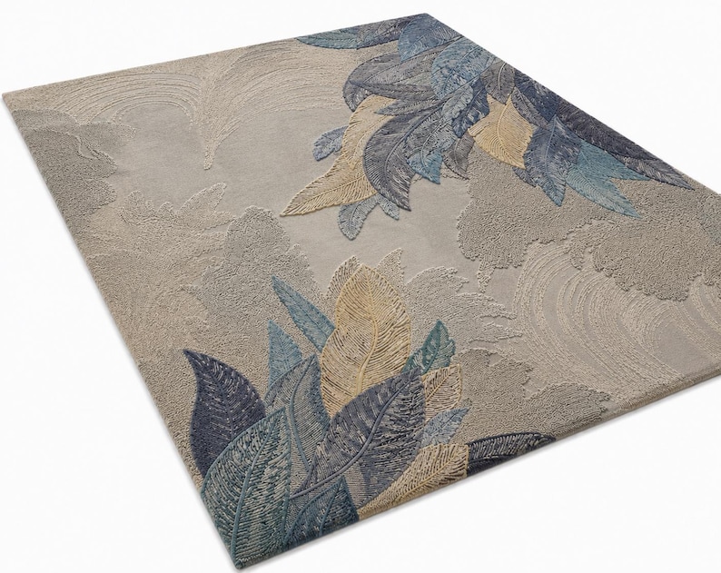 Op de afbeelding: Een rechthoekig vloerkleed met een beige achtergrond en een gedetailleerd bladerontwerp in tinten blauw, goud en groen. De bladeren zijn ingewikkeld patroon, waardoor een textuur ontstaat. Het ontwerp van het vloerkleed voegt een vleugje door de natuur ge&iuml;nspireerde elegantie toe.