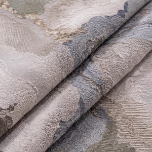 May include: A folded area rug featuring an abstract pattern with textured surfaces in shades of sage green, slate blue, cream, and beige. The design incorporates varying pile heights and organic shapes that create a layered, tactile appearance.