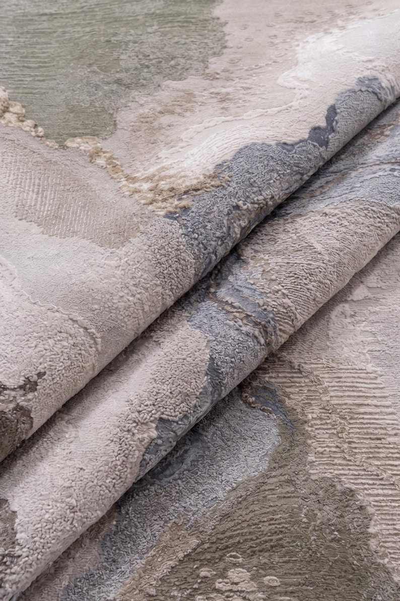 May include: A folded area rug featuring an abstract pattern with textured surfaces in shades of sage green, slate blue, cream, and beige. The design incorporates varying pile heights and organic shapes that create a layered, tactile appearance.