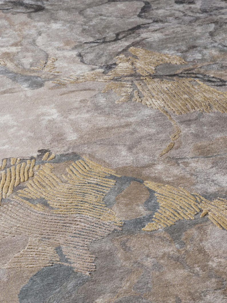 May include: A textured area rug featuring an abstract pattern in shades of beige, taupe, and charcoal gray. The design incorporates raised, carved-style golden-yellow sections with linear detailing that contrast against the smooth, mottled background.