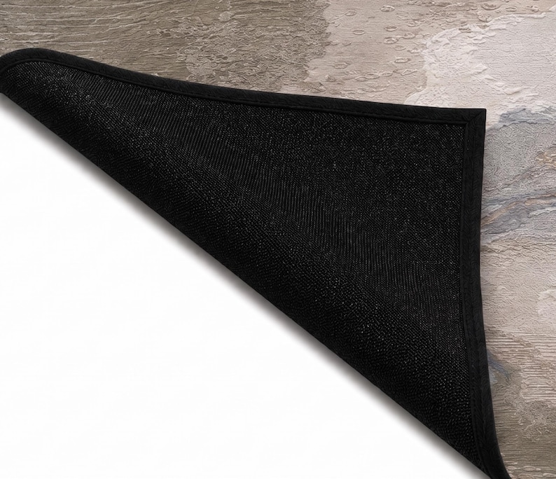 May include: A rectangular floor mat with a folded corner revealing a black, textured, non-slip underside. The top surface features a marbled pattern in shades of beige, gray, and white with a smooth finish and a solid black fabric border.
