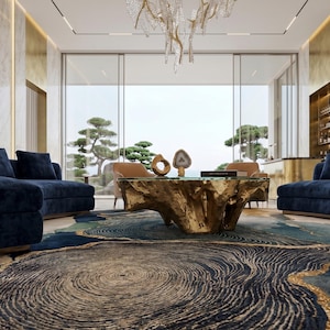 May include: A living room featuring a large, gold-finished, organic-shaped coffee table with a glass top, positioned on a textured rug with blue, gold, and cream concentric patterns. Two navy blue sofas flank the table, with a branch-like chandelier above.