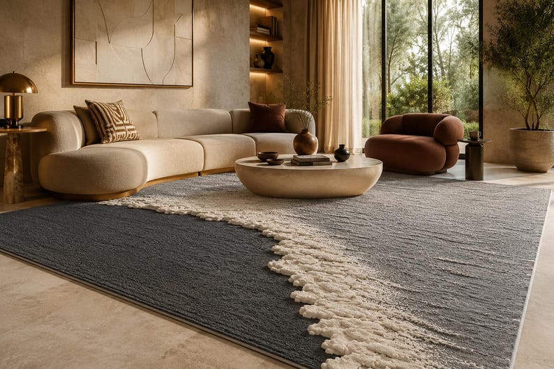 Op de afbeelding: Een moderne woonkamer met een groot, getextureerd vloerkleed met een golfachtig ontwerp in grijstinten en cr&egrave;me. De kamer omvat een gebogen beige bank, een ronde salontafel en een bruine fauteuil, wat een gezellige en stijlvolle sfeer cre&euml;ert.