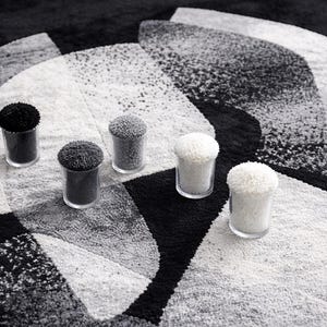 May include: A monochrome rug with a geometric pattern, featuring five small glass containers filled with textured material. The containers hold black, grey, and white materials. The rug's design is abstract.