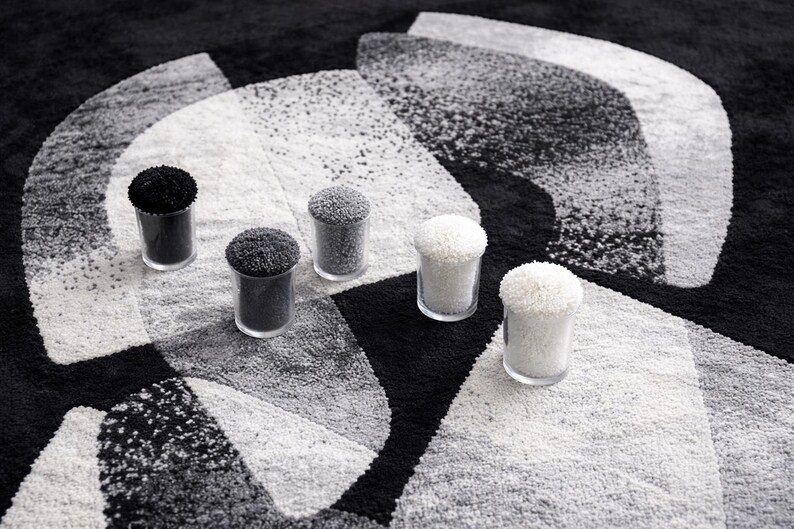 May include: A monochrome rug with a geometric pattern, featuring five small glass containers filled with textured material. The containers hold black, grey, and white materials. The rug's design is abstract.