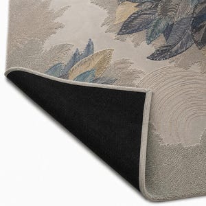 Op de afbeelding: Een rechthoekig tapijt met een gedetailleerd bloemmotief met blauwe, gouden en groene bladeren op een beige achtergrond. De rand van het tapijt is lichtbeige en de onderkant is zwart. Het ontwerp bevat textuurelementen.