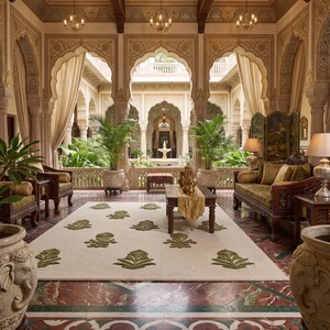 May include: A grand interior space featuring a cream-colored rug with green leaf patterns, a dark wood coffee table with a gold runner, and two large stone planters carved with elephant heads. The room has marble floors and arched doorways.