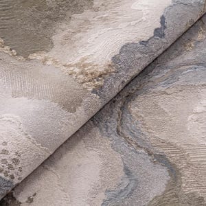 May include: A folded area rug featuring an abstract pattern with textured layers in shades of beige, taupe, cream, and slate blue. The surface displays varied pile heights and intricate woven details that create a multi-dimensional appearance.