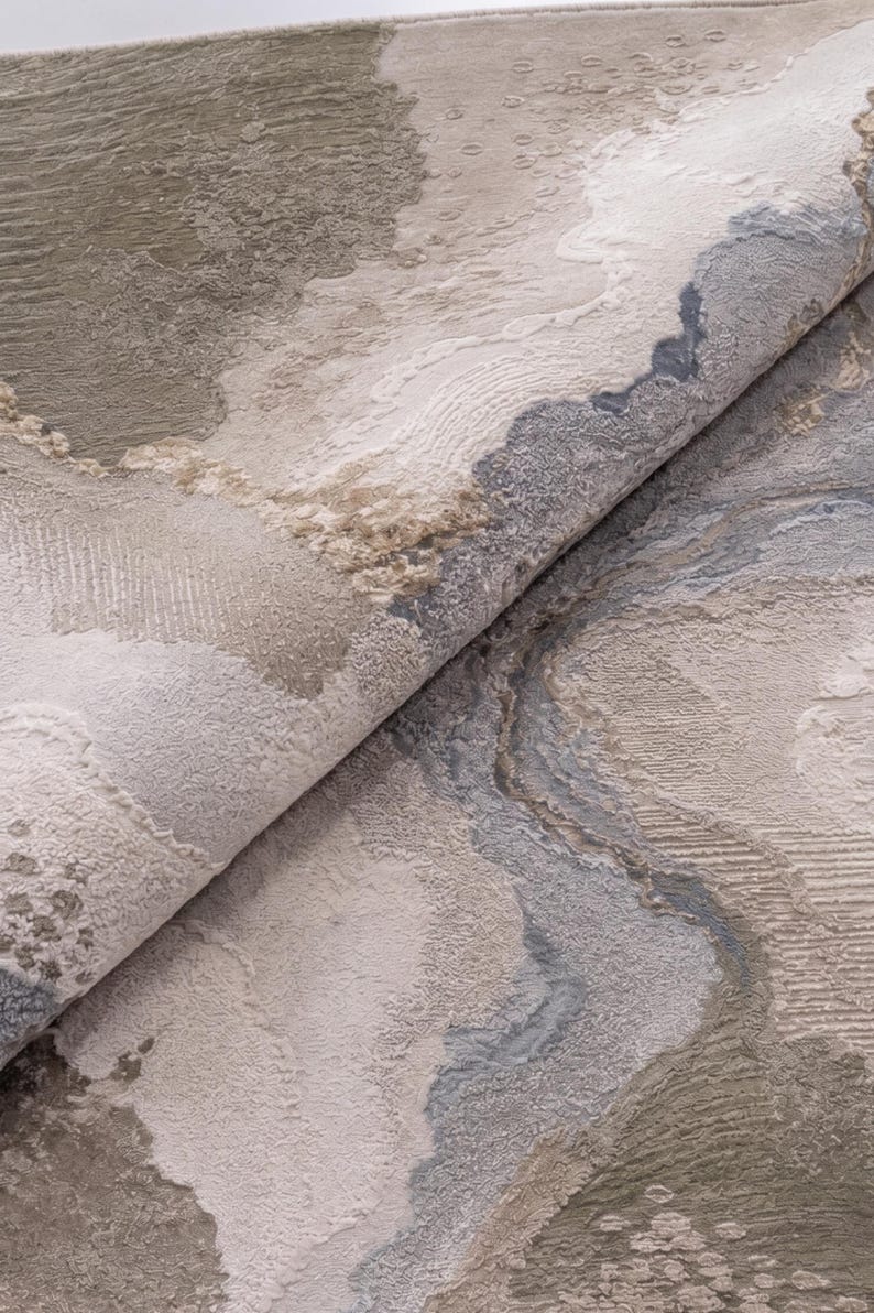 May include: A folded area rug featuring an abstract pattern with textured layers in shades of beige, taupe, cream, and slate blue. The surface displays varied pile heights and intricate woven details that create a multi-dimensional appearance.