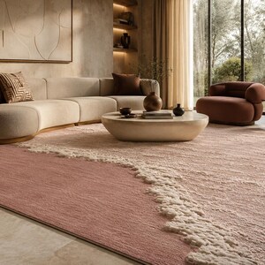 Op de afbeelding: Een moderne woonkamer met een groot, roze vloerkleed met een textuurontwerp. Het kleed heeft een golfachtig patroon in wit langs &eacute;&eacute;n rand. Een beige hoekbank, een ronde salontafel en een bruine fauteuil zijn ook zichtbaar.