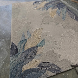 Op de afbeelding: Een groot, rechthoekig vloerkleed met een gedetailleerd veermotief. Het vloerkleed heeft een neutrale beige achtergrond met verhoogde, getextureerde patronen van gestileerde veren in tinten blauw, grijs en bruin. Het ontwerp bedekt een groot deel van het oppervlak van het vloerkleed.