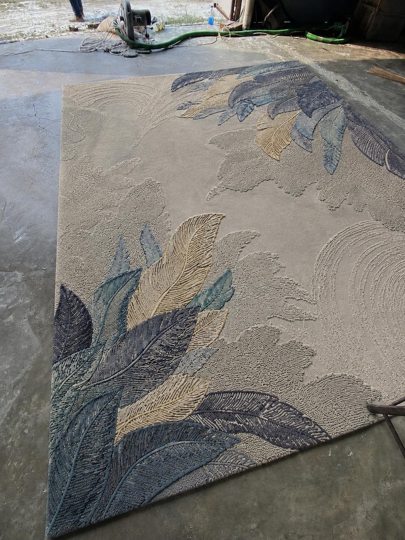 Op de afbeelding: Een groot, rechthoekig vloerkleed met een gedetailleerd veermotief. Het vloerkleed heeft een neutrale beige achtergrond met verhoogde, getextureerde patronen van gestileerde veren in tinten blauw, grijs en bruin. Het ontwerp bedekt een groot deel van het oppervlak van het vloerkleed.