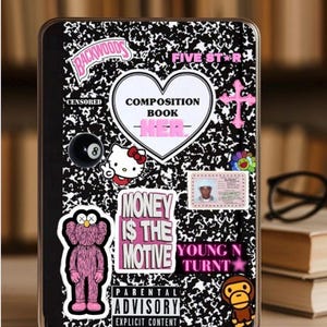 May include: A black and white composition notebook phone case with various stickers. Stickers include a pink cross, a Hello Kitty, a black 8-ball, and text that reads "MONEY IS THE MOTIVE" and "PARENTAL ADVISORY EXPLICIT CONTENT".