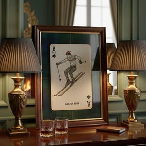 May include: A framed playing card featuring a skier, labeled "ACE OF SKIS." The card is set against a plaid background within a dark wooden frame. Two ornate lamps with pleated shades flank the frame on a dark wood table.