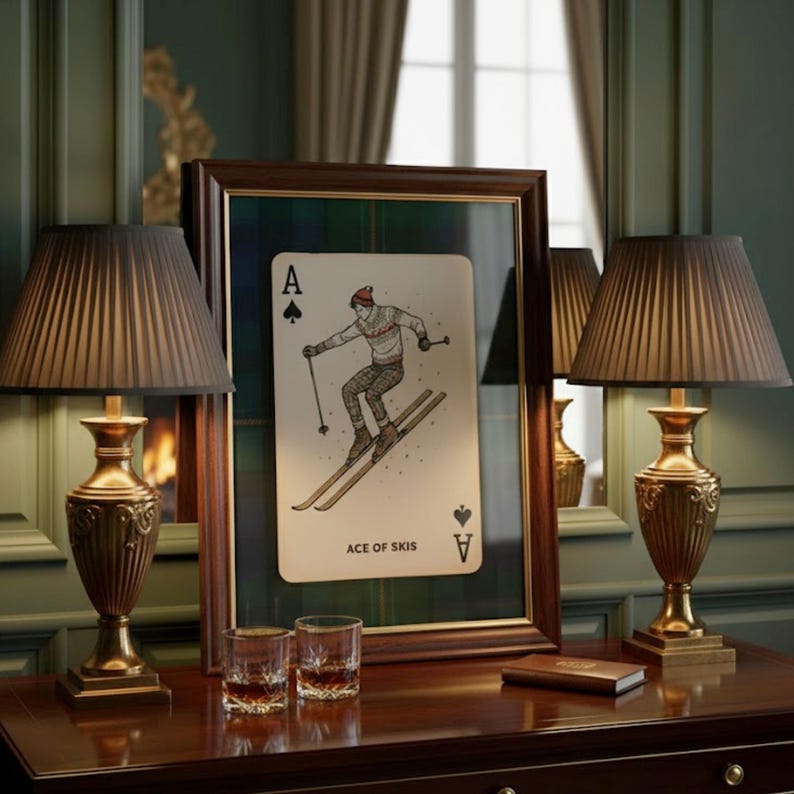 May include: A framed playing card featuring a skier, labeled "ACE OF SKIS." The card is set against a plaid background within a dark wooden frame. Two ornate lamps with pleated shades flank the frame on a dark wood table.