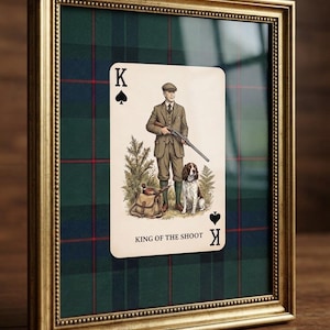 Pode incluir: Uma impressão de carta de jogar emoldurada com uma pessoa em fato de tweed segurando uma espingarda, acompanhada por um cão de caça e um faisão. A carta está rotulada como King of the Shoot sobre um fundo xadrez verde, azul e vermelho.