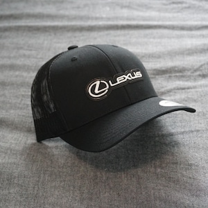 May include: Black baseball cap with a mesh back and a curved brim. The cap features the Lexus logo in white on a black rectangular patch on the front. The hat is set against a gray background.