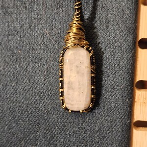 May include: A handcrafted pendant featuring a rectangular, light-colored stone encased in a wire-wrapped frame. The frame is a combination of black and gold-colored wire, suspended from a brown cord. The pendant is displayed against a denim background.