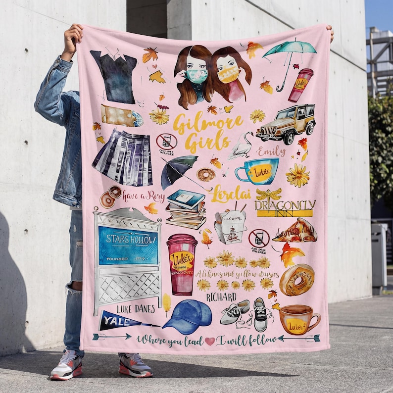 May include: Pink blanket featuring illustrations and text related to the TV show "Gilmore Girls." Includes images of characters, coffee cups, a car, and the text "Gilmore Girls," "Luke's," and "Stars Hollow."