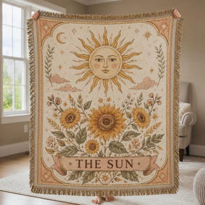 May include: A woven throw blanket featuring a central sun face illustration with rays, surrounded by sunflowers, foliage, and a crescent moon. The bottom includes a banner with the text THE SUN. The blanket has a fringed edge in beige and gold.