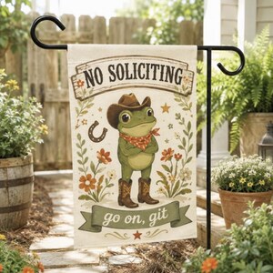 May include: A rectangular garden flag featuring a green frog wearing a brown cowboy hat, a red patterned bandana, and brown cowboy boots. The text NO SOLICITING is printed at the top, and the text go on, git is printed on a green banner below.