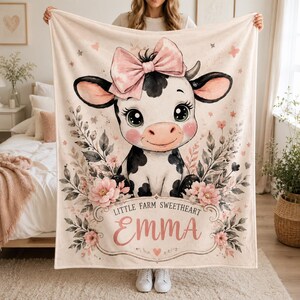 May include: A cream-colored blanket featuring a cartoon cow with a pink bow, black and white spots, and floral accents. The text "Little Farm Sweetheart Emma" is displayed in a decorative frame. The blanket is held up by a person.