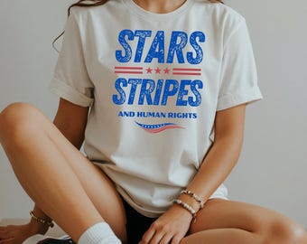 May include: Cream-colored t-shirt with the words "STARS STRIPES AND HUMAN RIGHTS" in blue. The text is arranged in a patriotic design with stars and stripes. The shirt has short sleeves and a relaxed fit.