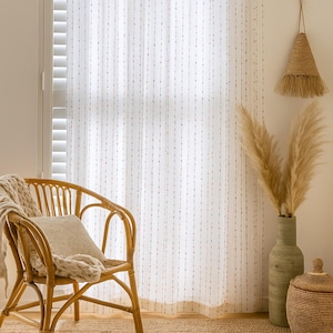 Puede incluir: Escena interior luminosa con una silla de rat&aacute;n tejido, un coj&iacute;n y una manta color crema. Cortinas blancas transparentes con patrones de puntos verticales enmarcan la ventana. Una l&aacute;mpara tejida colgante y un jarr&oacute;n con hierbas secas completan la escena.