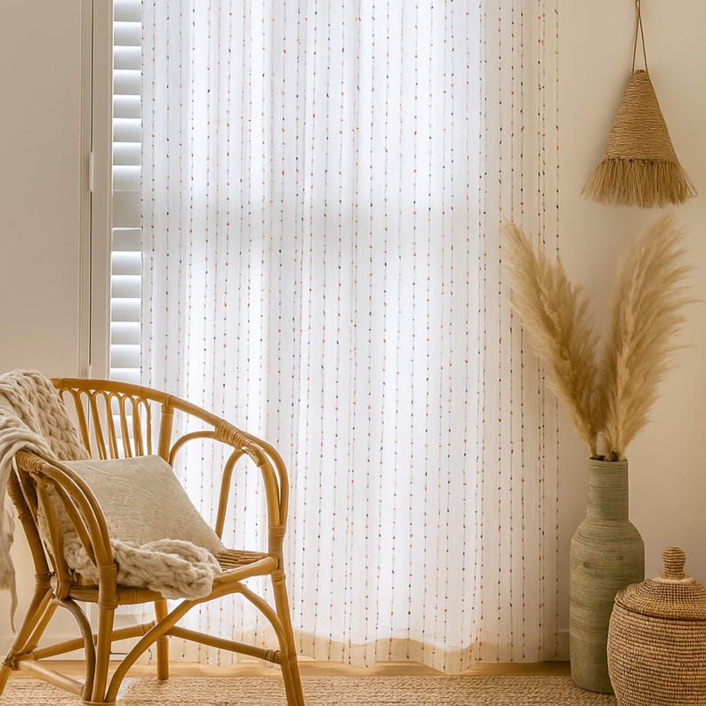 Puede incluir: Escena interior luminosa con una silla de rat&aacute;n tejido, un coj&iacute;n y una manta color crema. Cortinas blancas transparentes con patrones de puntos verticales enmarcan la ventana. Una l&aacute;mpara tejida colgante y un jarr&oacute;n con hierbas secas completan la escena.