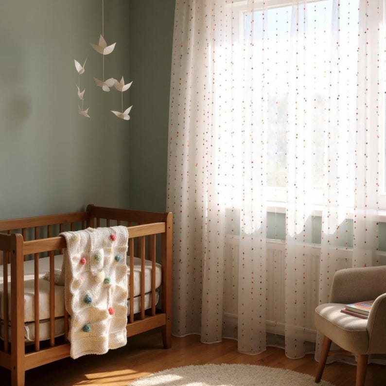 Puede incluir: Una habitaci&oacute;n infantil con una cuna de madera, una manta crema con pompones de colores y cortinas transparentes con lunares rojos y rosas. Un m&oacute;vil de p&aacute;jaros de papel cuelga sobre la cuna. Un sill&oacute;n beige est&aacute; cerca de la ventana.