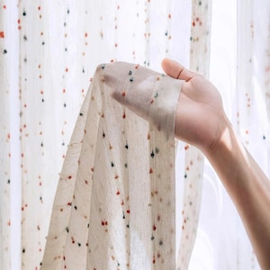 Puede incluir: Tela de cortina transparente blanquecina con rayas verticales y peque&ntilde;os pompones de colores. La tela se sostiene, mostrando su transparencia. Los pompones son en tonos rojos, verdes y marrones. Ideal para la decoraci&oacute;n del hogar.