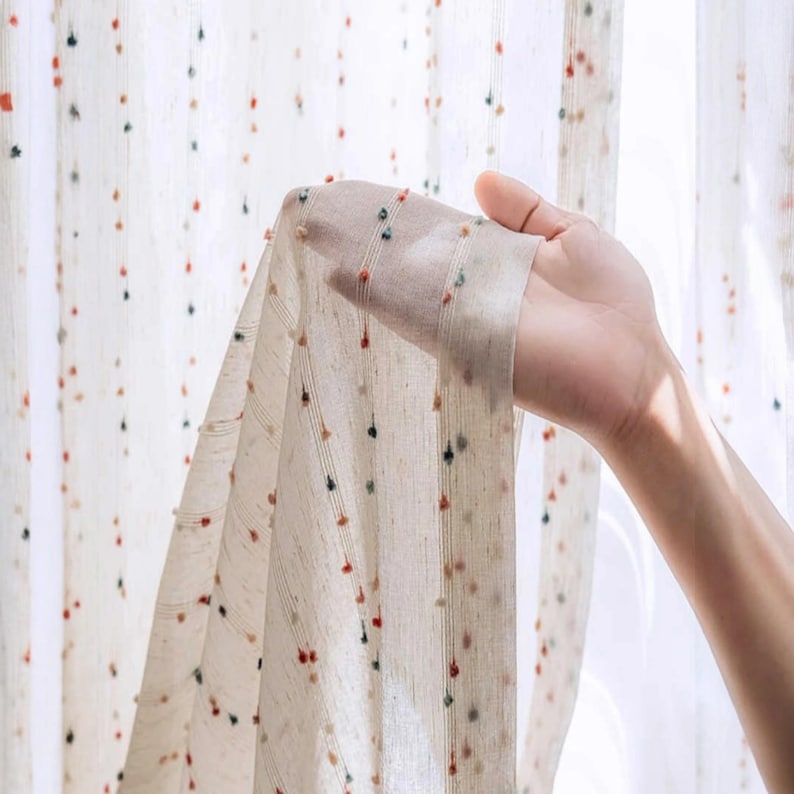 Puede incluir: Tela de cortina transparente blanquecina con rayas verticales y peque&ntilde;os pompones de colores. La tela se sostiene, mostrando su transparencia. Los pompones son en tonos rojos, verdes y marrones. Ideal para la decoraci&oacute;n del hogar.