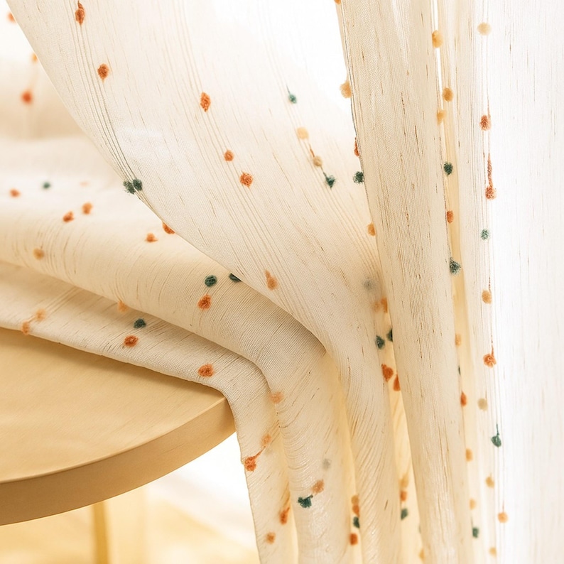 Puede incluir: Cortina transparente de color crema con una textura delicada y peque&ntilde;os pompones dispersos en naranja y verde. La cortina est&aacute; drapeada sobre una mesa de madera, creando pliegues suaves y permitiendo que la luz se filtre. Un tratamiento de ventana decorativo.