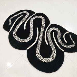 Puede incluir: Alfombra negra con diseño de serpiente blanca. La alfombra presenta un gran patrón de serpiente abstracto con un patrón blanco texturizado sobre un fondo negro. El diseño de la serpiente está delineado en blanco.