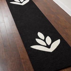 May include: Black rectangular rug with a white floral design. The rug is placed on a dark wood floor. The floral design is repeated at both ends of the rug.