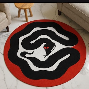 Può includere: Un tappeto rotondo con un design audace con un serpente nero su sfondo bianco, incorniciato da un bordo rosso vibrante. Gli occhi del serpente sono rossi. Il tappeto è su un pavimento di marmo.