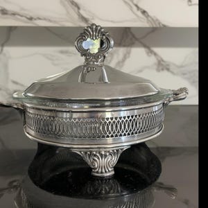 May include: A silver-plated serving dish with a glass lid. The dish features ornate detailing, including a decorative handle on the lid and a lattice-patterned band around the base. The dish is supported by three decorative feet.