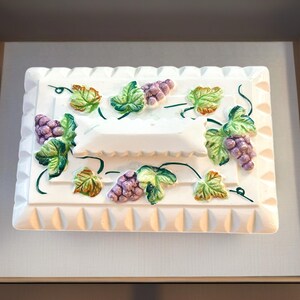 May include: A white rectangular ceramic dish with raised grape and leaf designs. The grapes are purple, and the leaves are green and yellow. The dish has a textured border and a central recessed area.