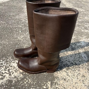 Puede incluir: Un par de botas altas de cuero marrón. Las botas tienen una puntera cuadrada y un puño doblado. Están hechas de cuero liso, marrón oscuro. Las botas están sobre una superficie de hormigón.