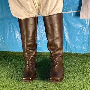 May include: A pair of brown leather knee-high boots with a square toe. The boots are made of a smooth, shiny leather and have a slight heel. The boots are worn with white pants.
