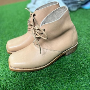 May include: A pair of light tan leather ankle boots with square toes and lace-up closures. The boots are set against a green background, showcasing their neutral color and classic design. The boots have a low heel.