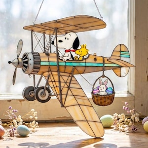 May include: Stained glass biplane featuring Snoopy and Woodstock. The plane has a brown and teal color scheme, with a basket of Easter eggs hanging below. The design is suitable for Easter decor.