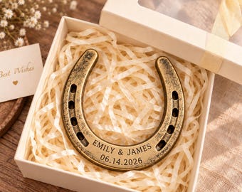 May include: A bronze-toned metal horseshoe engraved with the names EMILY & JAMES and the date 06.14.2026, resting inside a white gift box filled with shredded paper filler, accompanied by a small card with the text Best Wishes.