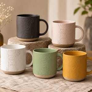 May include: Five ceramic mugs in black, light pink, white, sage green, and mustard yellow. Each mug features an embossed tree trunk design with a heart carving containing the text C and A and the date 10-21-16. The mugs have a smooth handle and a contrasting unglazed tan base.
