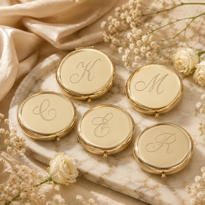 May include: Five round gold-toned metal compact mirrors, each featuring a unique engraved cursive letter on the lid. The letters K, M, C, E, and R are centered on the polished surfaces of these circular cases, which are arranged together.