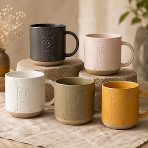 May include: Five ceramic mugs in black, light pink, white, olive green, and mustard yellow. Each mug features an embossed tree trunk design with a heart containing the initials J+E and the date 1.15.23. The mugs have a smooth finish and a base.