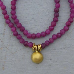 May include: A double-strand necklace featuring small, faceted, deep magenta-colored beads. The necklace includes a central gold-toned pendant shaped like a smooth, rounded disc, suspended from a series of small, vertical gold-toned metal rings.